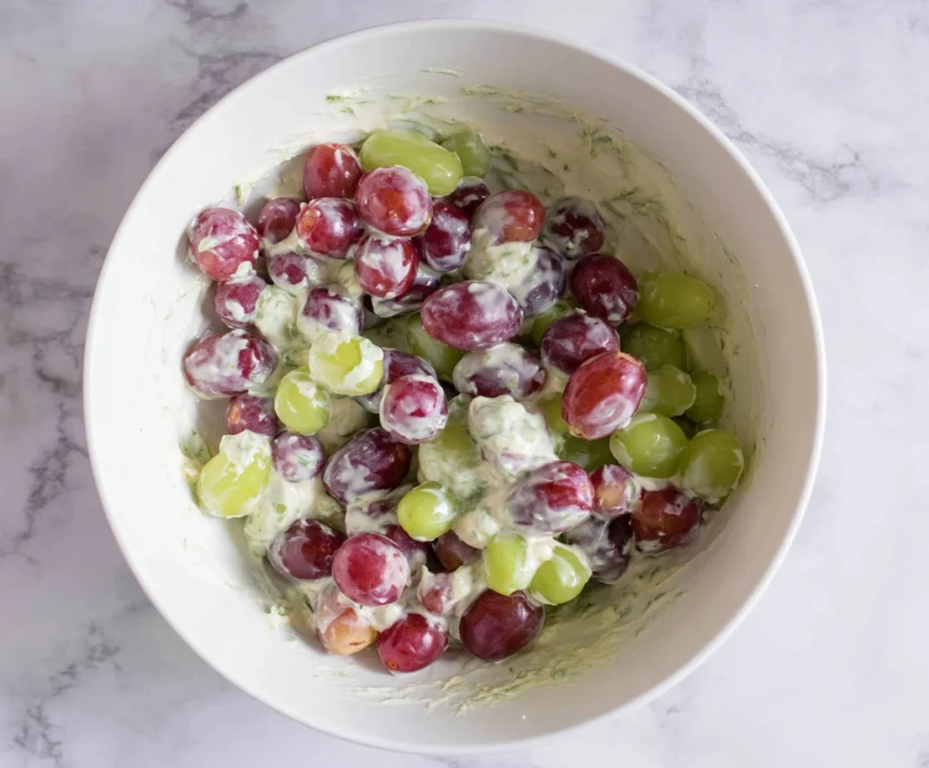 Gently folding red and green grapes into the creamy cream cheese dressing in a large bowl